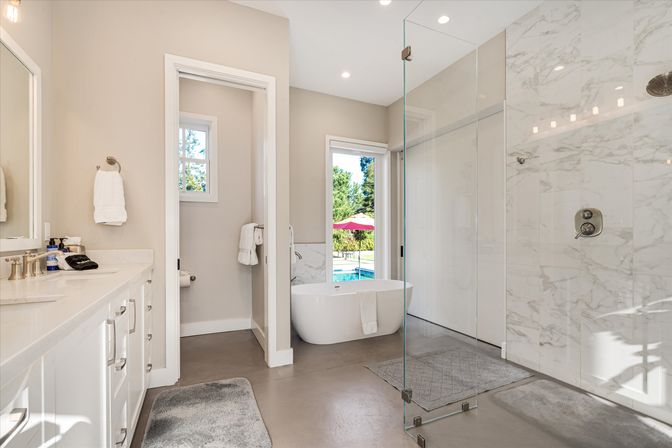 Bright modern bathroom with double vanity, freestanding tub by glass door overlooking a backyard pool and umbrella, and a glass walk-in shower with marble tile.