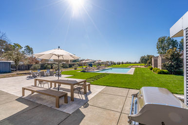 Sun-splashed backyard patio with wooden picnic table and umbrella, stainless steel grill, manicured green lawn leading to a rectangular swimming pool with lounge chairs under a clear blue sky.