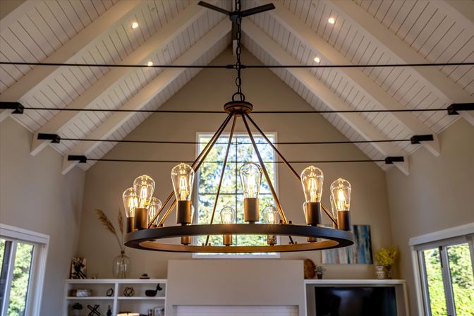 Warm-glow round metal chandelier with exposed Edison bulbs hangs in a sunlit living room with vaulted white beamed ceiling, fireplace and built-in shelves.
