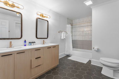 Bright, modern spa-inspired bathroom with light wood double-sink vanity, brass mirrors and faucets, dark hexagonal floor tiles, and a tub-shower with vertical neutral tiles and white curtain.