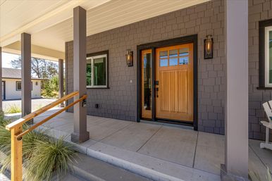 Covered suburban front porch with natural wood paneled front door and glass sidelight, gray shingle siding, concrete steps, wooden handrail, and black wall lanterns