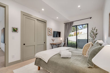 Sunlit modern bedroom with sage-green quilted bed and pillows, folded towels on the footboard, sliding closet doors, wall-mounted TV and small desk, gray curtains framing a glass patio door overlooking a private backyard.