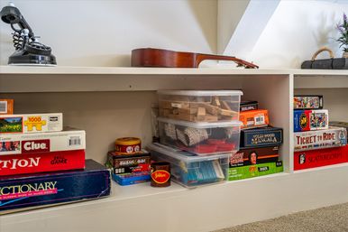 Home playroom shelf packed for game night with stacked board games, clear plastic bins of game pieces, a ukulele resting on the top shelf and a retro rotary phone