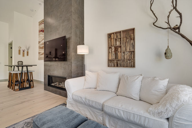 Contemporary open-concept living room with white slipcovered sofa and fluffy throw, blue ottoman, wall-mounted TV above a gray tile fireplace, carved wooden wall art and hanging branch decor, light hardwood floors and a breakfast bar in the background.