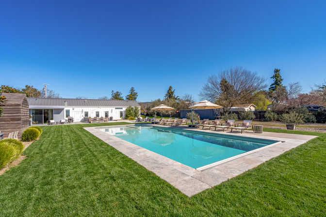 Sunlit suburban backyard with a rectangular outdoor swimming pool and clear turquoise water, stone deck lined with lounge chairs and umbrellas, manicured green lawn and modern white pool house under a bright blue sky.