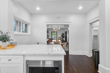 Bright open-concept kitchen with white marble island and built-in microwave, dark hardwood floors, potted plant on counter, looking through wide doorway to a dining table and living room with a modern chandelier.