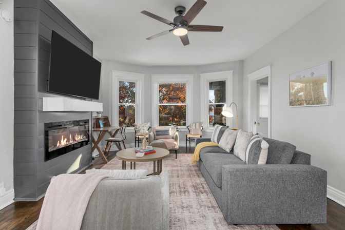 Cozy modern living room with gray sectional and armchair, round coffee table, wall-mounted TV above a linear gas fireplace, bay windows showing autumn foliage, ceiling fan, and hardwood floors.