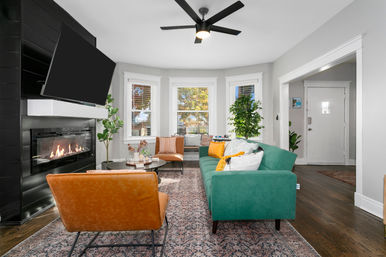 Bright modern living room with green sofa, tan leather chairs, gas fireplace under a wall TV, bay windows, potted plants, patterned rug and ceiling fan