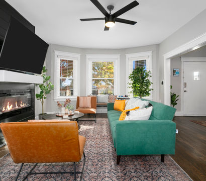 Bright modern living room with green sofa, tan leather chairs, gas fireplace under a wall TV, bay windows, potted plants, patterned rug and ceiling fan