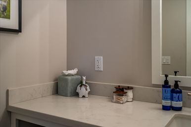 Modern bathroom vanity marble countertop with green tissue box, small white ceramic animal figurine, glass jars of cotton swabs and pads, blue soap and lotion bottles, mirror and wall outlet.