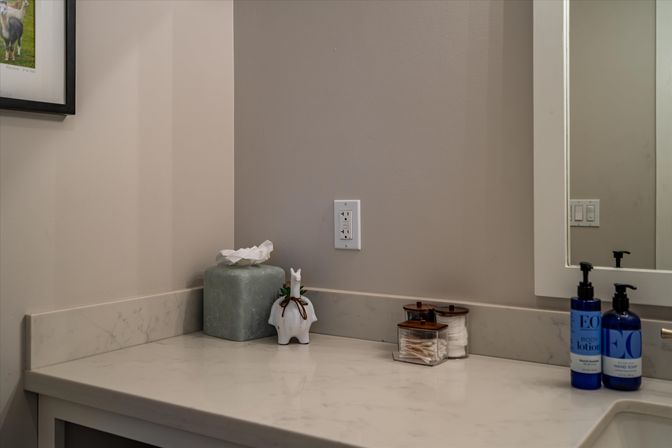 Modern bathroom vanity marble countertop with green tissue box, small white ceramic animal figurine, glass jars of cotton swabs and pads, blue soap and lotion bottles, mirror and wall outlet.