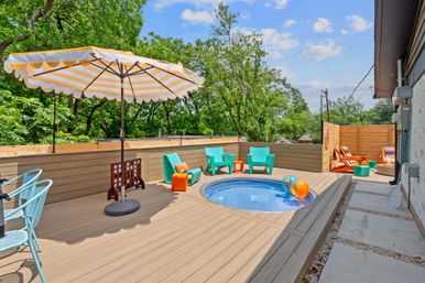 Sunny rooftop deck with round plunge pool, yellow-striped umbrella, turquoise lounge chairs, colorful beach balls and cozy Adirondack seating on wood decking surrounded by trees.