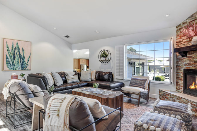 Sunlit suburban living room with dark leather sectional, wooden coffee table, stone fireplace with glowing fire, large paned window overlooking a patio, neutral pillows and patterned poufs in modern-rustic style.