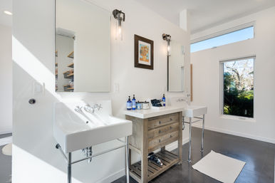 Sunlit modern bathroom with two white pedestal sinks, rustic wood vanity with drawers, matching mirrors and wall sconces, and a large window revealing a green garden view.