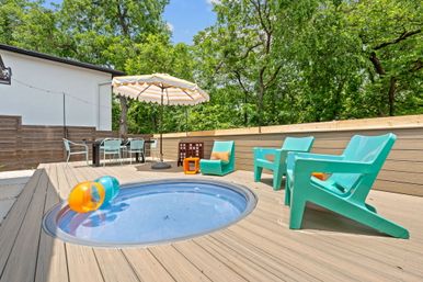 Sunny backyard wooden deck with a small circular plunge pool, colorful inflatable balls, aqua Adirondack-style chairs, striped patio umbrella, outdoor dining table and lush green trees