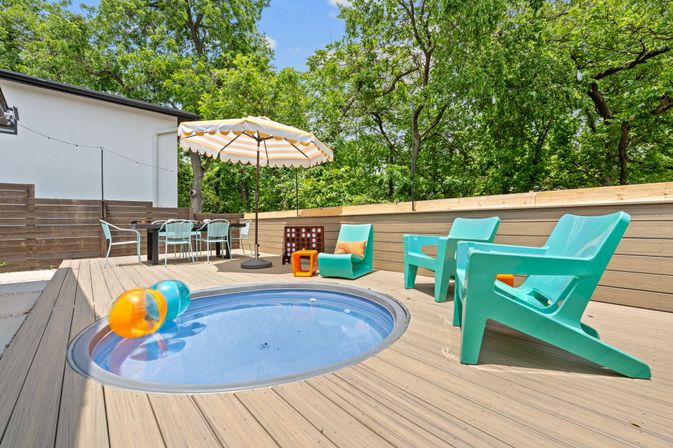 Sunny backyard wooden deck with a small circular plunge pool, colorful inflatable balls, aqua Adirondack-style chairs, striped patio umbrella, outdoor dining table and lush green trees