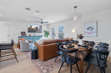 Bright modern open-concept living and dining room with tan leather sofa, textured armchair, blue sideboard, wall-mounted TV showing a city skyline, wooden dining table with black chairs set for dinner, patterned rug, pendant light, and playful armadillo wall art reading Rolling with the Homies.