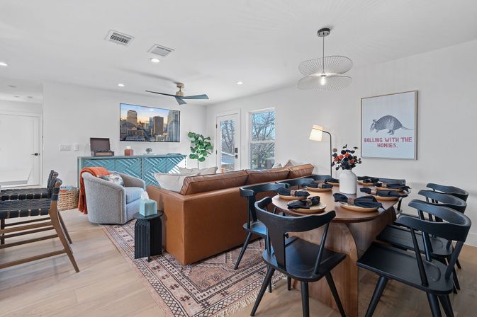 Bright modern open-concept living and dining room with tan leather sofa, textured armchair, blue sideboard, wall-mounted TV showing a city skyline, wooden dining table with black chairs set for dinner, patterned rug, pendant light, and playful armadillo wall art reading Rolling with the Homies.