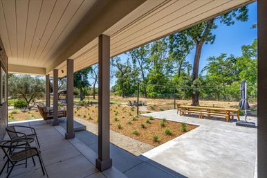 Sunny covered backyard porch with chairs and cozy seating, concrete patio leading to a picnic-style dining bench, mulched planting beds and mature trees under a blue sky.