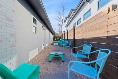 Narrow outdoor courtyard between modern homes with bright turquoise metal chairs, playful cornhole boards on a gravel path and a wooden privacy fence under a clear blue sky.