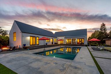 Modern farmhouse-style backyard at sunset with glowing interiors reflected in a rectangular pool, concrete patio, red umbrellas, lounge chairs, and a fire pit.