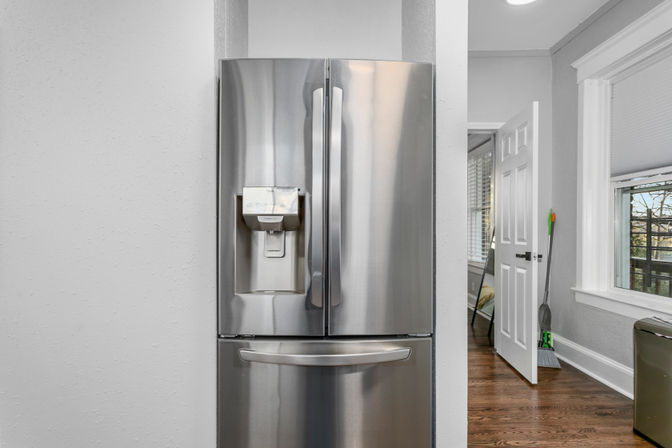 Sleek stainless-steel French-door refrigerator with water dispenser and bottom freezer drawer in a light-gray home kitchen with hardwood floors, open door to a hallway and a window view.