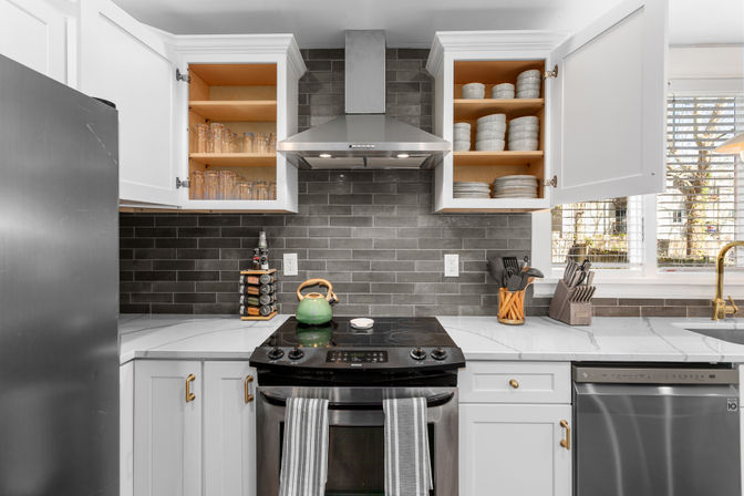 Bright modern home kitchen with gray subway-tile backsplash, white cabinets with open shelving showing dishes and glasses, stainless steel range hood and dishwasher, black glass cooktop with green kettle, marble-look countertops and gold hardware under a sunlit window.