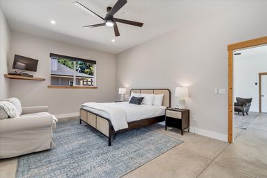 Bright modern bedroom with neutral walls, wooden-framed bed with white linens, matching nightstands and lamps, blue patterned area rug, ceiling fan, armchair and wall-mounted TV by a window overlooking neighboring home.