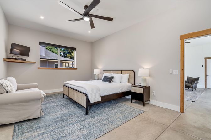 Bright modern bedroom with neutral walls, wooden-framed bed with white linens, matching nightstands and lamps, blue patterned area rug, ceiling fan, armchair and wall-mounted TV by a window overlooking neighboring home.