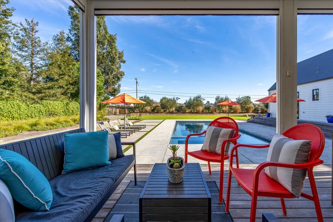 Sunny backyard pool patio with rectangular swimming pool, modern outdoor seating — navy sofa with turquoise cushions, two red metal chairs, coffee table with potted plant, lounge chairs and orange umbrellas, green hedges and blue sky