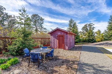 Cozy backyard scene with a red garden shed, round patio table with blue cushioned chairs, gravel path and evergreen trees under a bright blue sky.