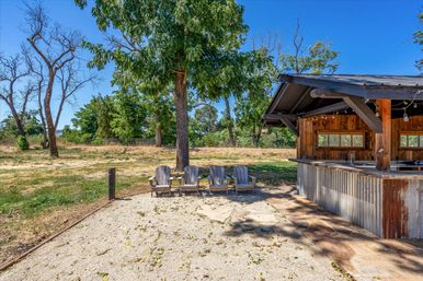 Rustic outdoor bar pavilion with corrugated metal counter and string lights, four Adirondack chairs on a gravel patio under a large shade tree in a sunny countryside yard