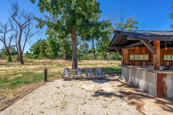 Rustic outdoor bar pavilion with corrugated metal counter and string lights, four Adirondack chairs on a gravel patio under a large shade tree in a sunny countryside yard