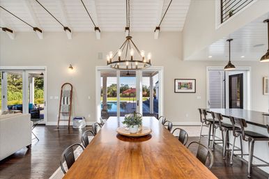 Sunlit open-concept dining room with long wooden table, metal chairs and farmhouse chandelier, sliding glass doors framing a backyard pool and patio, adjacent kitchen island with bar stools and vaulted white-beamed ceiling.