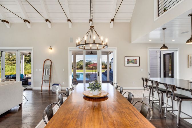 Sunlit open-concept dining room with long wooden table, metal chairs and farmhouse chandelier, sliding glass doors framing a backyard pool and patio, adjacent kitchen island with bar stools and vaulted white-beamed ceiling.