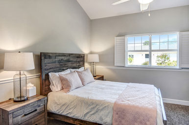 Cozy sunlit bedroom with rustic wood bedframe, white linens and blush-pink quilt and pillows, matching nightstands with glass lamps, and a shuttered window letting in daylight.