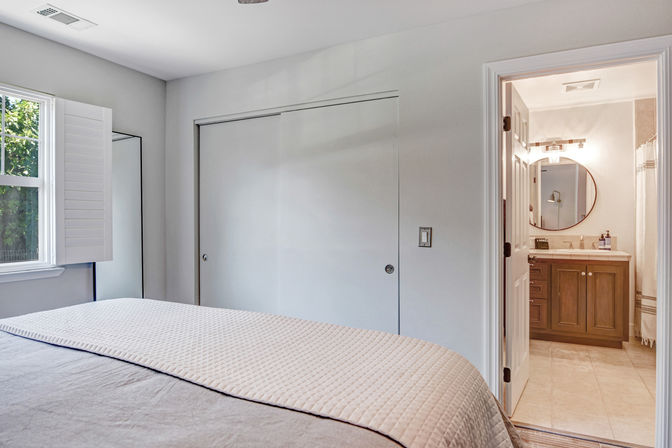 Bright master bedroom with quilted bedspread, shuttered window, sliding closet doors, and open doorway to ensuite bathroom with wooden vanity and round mirror.