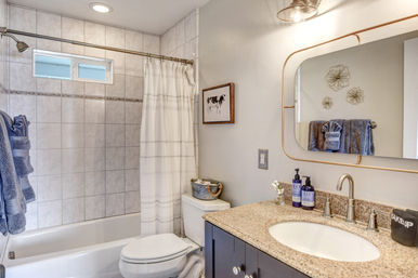 Bright contemporary home bathroom with white tiled tub and shower, striped curtain, cute cow wall art, granite vanity with oval mirror and bronze faucet, and rolled blue towels.