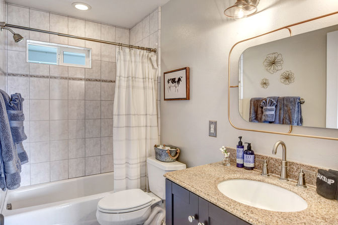 Bright contemporary home bathroom with white tiled tub and shower, striped curtain, cute cow wall art, granite vanity with oval mirror and bronze faucet, and rolled blue towels.