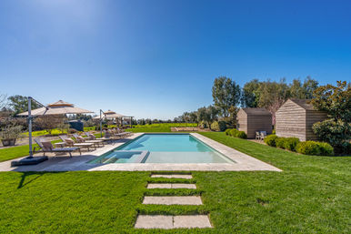 Resort-style outdoor swimming pool in a landscaped backyard with a stone path, sun loungers and umbrellas, wooden cabanas, manicured green lawn and clear blue sky.