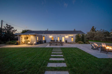 Twilight view of a modern farmhouse backyard with a green lawn and stepping-stone path leading to a lit patio, outdoor dining table, cozy lounge area and fire pit.