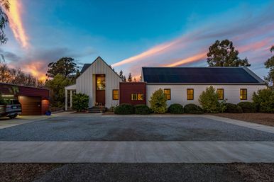 Modern farmhouse-style home with solar-panel roof, warm glowing windows, gravel driveway and detached garage at vibrant sunset, framed by trees and neatly trimmed shrubs.