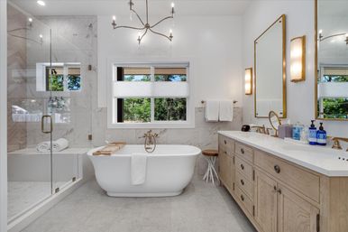 Bright spa-like residential bathroom with a freestanding white tub, brass floor-mount faucet and wooden tub tray, glass-enclosed marble shower, large window with tree views, double vanity with wood cabinets, brass mirrors and sconces, and neutral tile flooring.