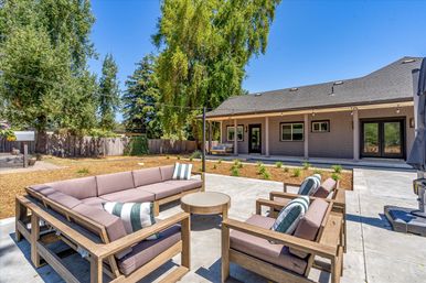 Sunny suburban backyard patio with wooden sectional sofa and armchairs around a round coffee table on a concrete slab, covered porch of a single-story house and large shade trees under a clear blue sky.