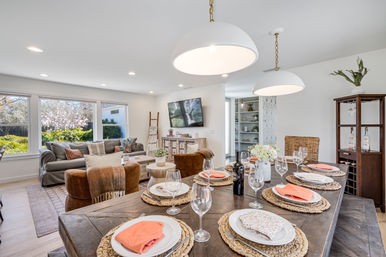 Bright open-plan dining and living room with rustic wooden table set for six (coral napkins, woven placemats, wine glasses), oversized white pendant lights, cozy gray sofa and brown chairs, wall-mounted TV, and large windows with a sunny garden view.