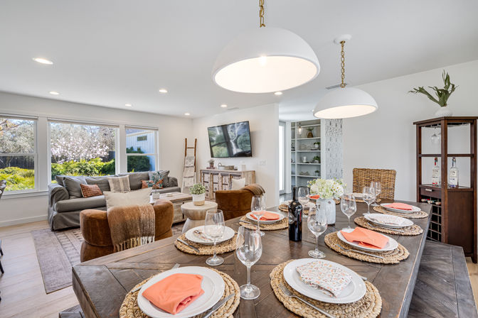 Bright open-plan dining and living room with rustic wooden table set for six (coral napkins, woven placemats, wine glasses), oversized white pendant lights, cozy gray sofa and brown chairs, wall-mounted TV, and large windows with a sunny garden view.