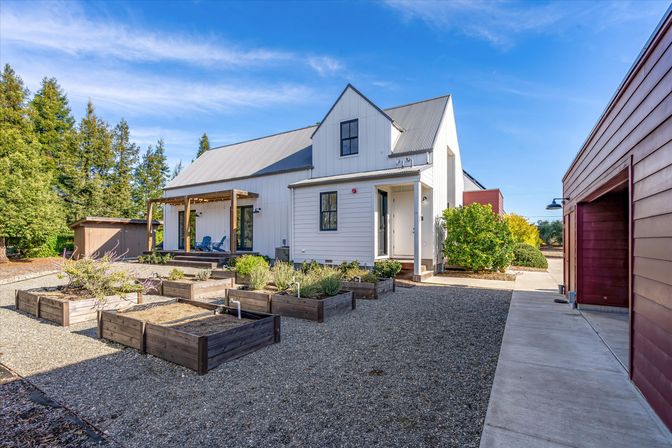 Modern white farmhouse with gray metal roof and covered wooden pergola, gravel courtyard with multiple raised wooden garden beds, cheery blue porch chairs, red detached garage to the right and evergreen trees under a bright blue sky