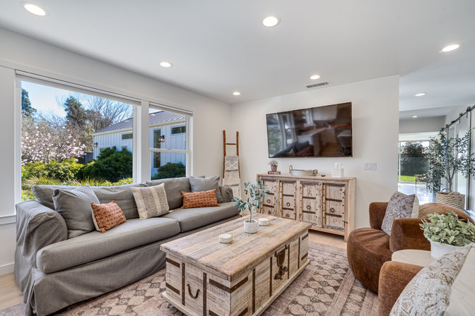 Sunlit modern farmhouse-style living room with large garden-facing windows, gray slipcovered sofa with patterned pillows, rustic wooden coffee table and matching sideboard, wall-mounted TV and cozy brown accent chairs.