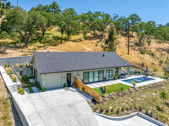 Modern single-story ranch-style home on an oak-studded hillside with a large concrete driveway, sliding glass doors to a patio, swimming pool and spa, and landscaped yard framed by dry grassy slopes.