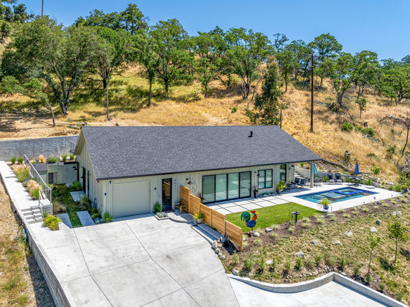 Modern single-story ranch-style home on an oak-studded hillside with a large concrete driveway, sliding glass doors to a patio, swimming pool and spa, and landscaped yard framed by dry grassy slopes.